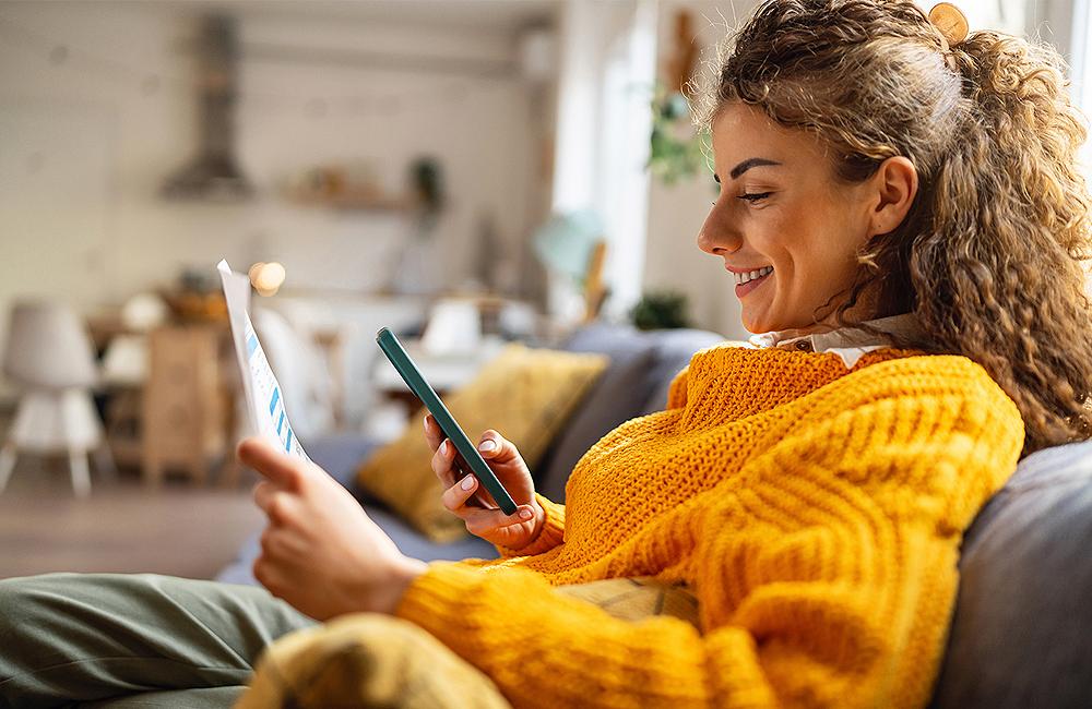 Jonge vrouw met gele trui kijkt lachend op haar smartphone in de zetel