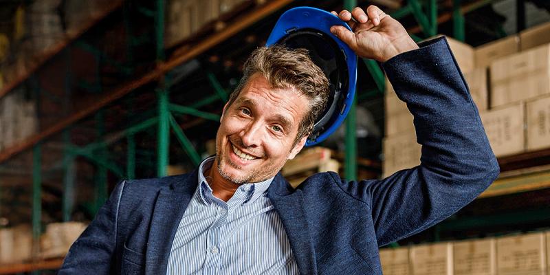 Een jonge zakenman staat vrolijk in zijn magazijn met veiligheidshelm