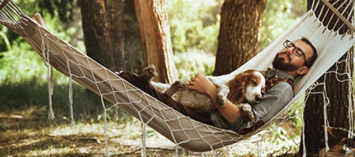 Man met baard en bril slaapt met zijn hond buiten in een hangmat 